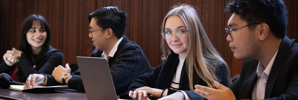 A group of University of Bristol Law students discussing work over laptops and papers at the wooden tables in the Old Council Chamber of the Wills Memorial Building.